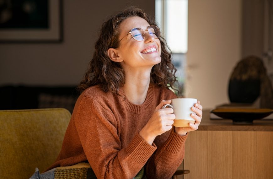 Young woman smiling while sitting on a couch at home and holding a cup filled with hot chocolate.