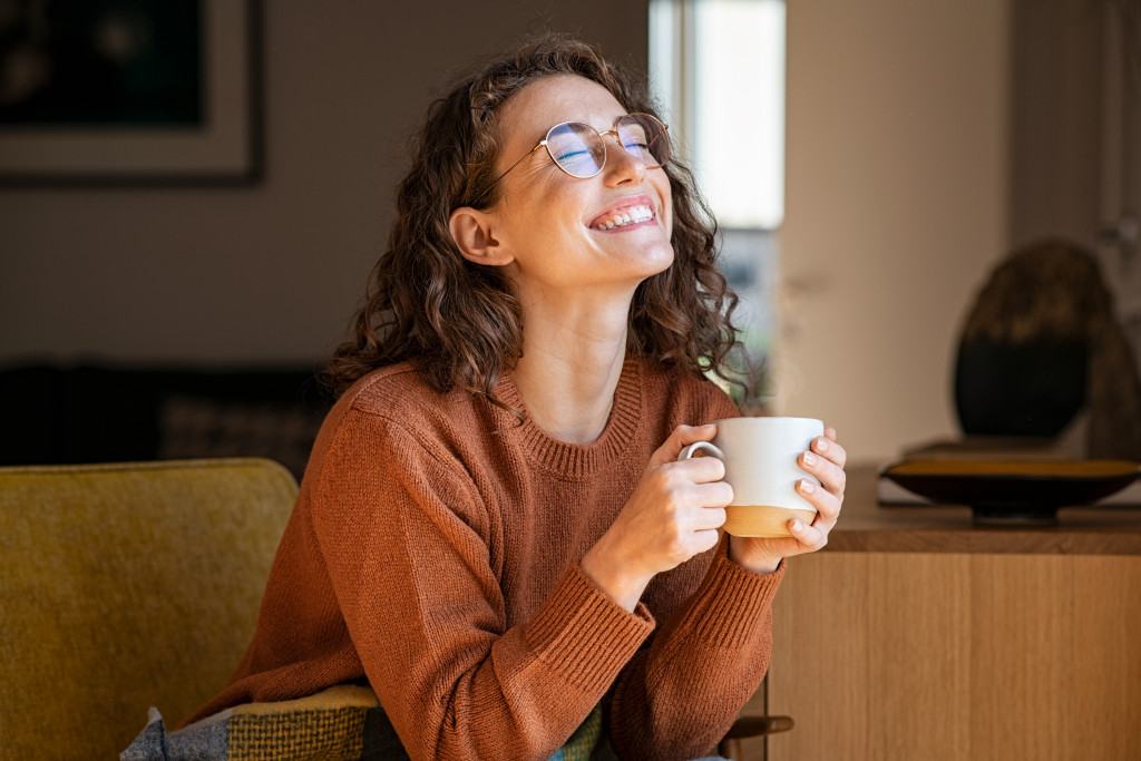 Young woman smiling while sitting on a couch at home and holding a cup filled with hot chocolate.